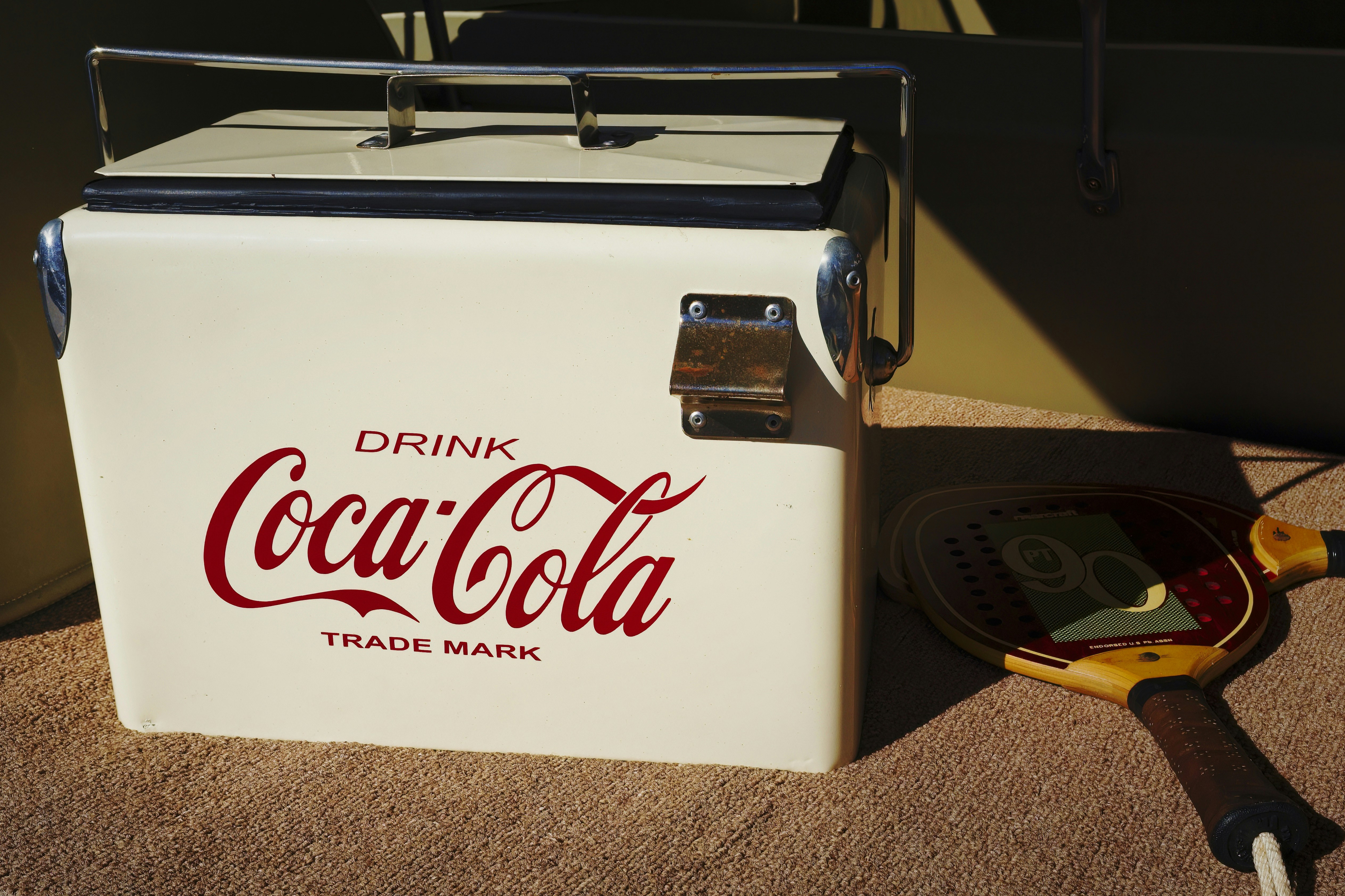 Vintage White Coca-Cola Cooler in a Car Trunk With a Paddle Racket - A classic white Coca-Cola cooler with a red logo sits in the back of a car. It is beside a paddle racket, suggesting a day of outdoor activities.